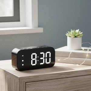 May include: A black digital alarm clock displaying 8:30 in white numerals. The clock sits on a wooden nightstand next to a small succulent in a white pot and a stack of books. Decorative string lights are draped across the books.