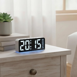 Op de afbeelding: Een digitale wekker met een witte lijst toont de tijd 20:15 in helderwitte cijfers. De klok toont ook de dag van de week. Hij staat op een lichtgekleurd houten nachtkastje, met een kleine plant en boeken in de buurt.