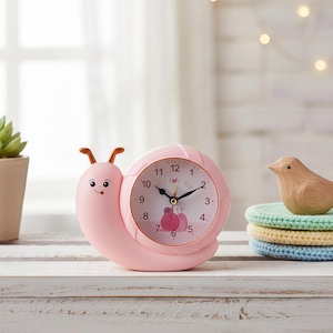 Può includere: Una sveglia rosa a forma di lumaca con un quadrante bianco. Il quadrante ha lancette nere e le parole "BEST FOR YOU". Un piccolo uccello di legno è appoggiato su una pila di sottobicchieri colorati.