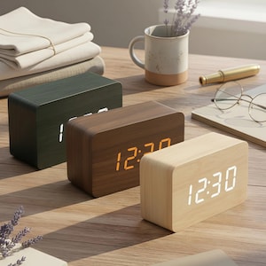 May include: Three rectangular wooden digital clocks in green, brown, and light wood tones, displaying the time. The clocks are on a wooden surface, with a mug, glasses, and a notebook in the background. The time displayed is 12:30.