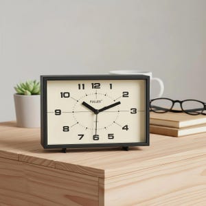Puede incluir: Un reloj analógico rectangular negro con una esfera crema, que muestra la hora. El reloj tiene números y manecillas negras, con la marca "FULLES" impresa en la esfera. Está sobre una superficie de madera, con una pequeña planta, libros y gafas en el fondo.