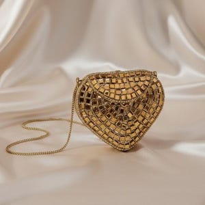 May include: A gold heart-shaped clutch bag with a chain strap. The bag is covered in small, square, reflective tiles, creating a mosaic effect. The background is a soft, cream-coloured fabric.