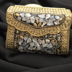 May include: A rectangular, gold-coloured clutch with detailed embossed patterns. The front is adorned with a mosaic of white and grey iridescent pieces, with gold-coloured leaf-shaped accents. The clasp is a simple, rectangular design.