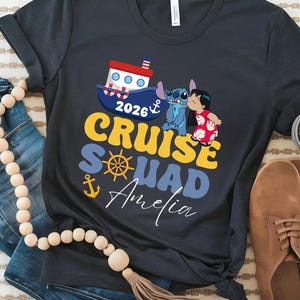 May include: Dark gray t-shirt with a colorful graphic that says "2026 Cruise Squad Amelia". The design includes a boat, anchor, and cartoon characters. The shirt is paired with jeans and brown shoes.
