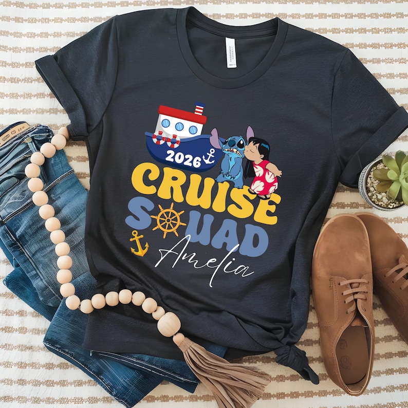 May include: Dark gray t-shirt with a colorful graphic that says "2026 Cruise Squad Amelia". The design includes a boat, anchor, and cartoon characters. The shirt is paired with jeans and brown shoes.
