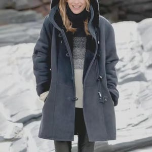 May include: A dark blue hooded coat with a toggle closure. The coat has a faux fur lining and a hood. The person in the image is wearing a black turtleneck sweater and a sweater with gray and white stripes.