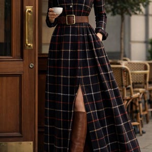 Puede incluir: Vestido largo a cuadros negros, rojos y crema, con cinturón marrón y botas altas a juego. El vestido tiene una parte delantera abotonada y mangas largas. Se ve un collar de cadena dorada.