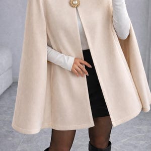 Op de afbeelding: Een crèmekleurige cape met een ronde halslijn en een decoratieve goudkleurige sluiting. De cape wordt gedragen over een wit shirt met lange mouwen en een zwarte rok. Zwarte laarzen maken de outfit compleet.