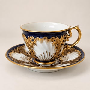 Peut inclure: Un ensemble tasse et soucoupe en porcelaine présentant un coloris bleu cobalt profond et blanc. L'extérieur de la tasse et la soucoupe sont ornés de motifs complexes en filigrane doré en relief et d'un design festonné en forme de coquille.