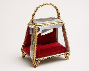 May include: A small display case with clear glass panels and a gold-toned metal frame. The interior base is lined with deep red velvet. It features a braided gold metal handle on top and stands on four ornate, decorative gold-toned metal feet.