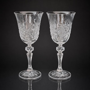 Puede incluir: Dos copas de vino de cristal transparente con intrincados patrones de corte en forma de diamante y abanico en los cálices. Cada copa tiene un tallo hexagonal facetado y una base circular con un diseño de estrella radiante.