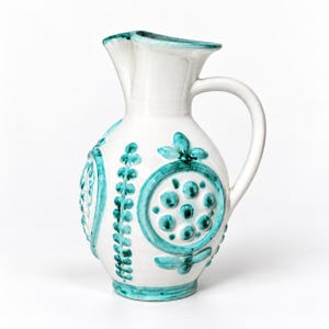Op de afbeelding: Een witte keramische kan met een ronde buik en een gebogen handvat. De buitenkant is versierd met reliëf, handgeschilderde blauwgroene bloem- en plantmotieven, waaronder cirkelvormige patronen en verticale bladvormen rondom de onderzijde.