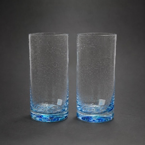 Puede incluir: Dos vasos cilíndricos altos hechos de vidrio transparente con una sutil textura de burbujas moteadas en todo el cuerpo. La base de cada vaso presenta una capa gruesa de vidrio azul vibrante con una densa concentración de pequeñas burbujas.