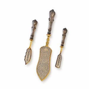 Könnte beinhalten: Ein Set aus drei Servierbestecken, bestehend aus einem Tortenheber und zwei kleineren Messern. Jedes Teil hat einen verzierten, silberfarbenen Griff mit geprägten Details und eine goldfarbene Klinge mit floralen Gravuren.