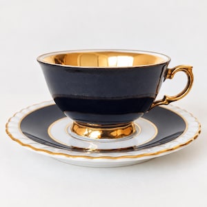 Op de afbeelding: Een theekop en schotel set met een diep marineblauwe buitenkant en een reflecterende metallic gouden binnenkant. De kop heeft een gouden handvat en voet, terwijl de bijpassende schotel een witte geschulpte rand heeft met een gouden afwerking.