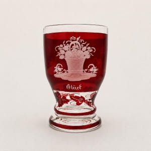 Pode incluir: Uma taça de vidro vermelho rubi com um design gravado de uma cesta cheia de flores e frutas. A palavra Glück está gravada em letras brancas abaixo da cesta. A base do copo é transparente com detalhes em vermelho e um padrão de folhas.