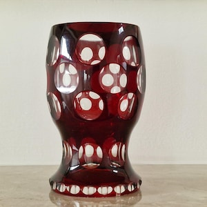 Op de afbeelding: Een robijnrode glazen vaas met een reeks witte stippen in het oppervlak. De vaas heeft een uitlopende basis en een afgeronde body, met een geschulpte rand aan de onderkant. De vaas is waarschijnlijk vintage of antiek.