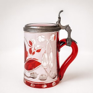 Op de afbeelding: Een bierpul van rood en helder geslepen glas met een bloemmotief en een metalen scharnierend deksel. De beker heeft een stevig rood glazen handvat en een cilindervormig lichaam met decoratieve gegraveerde blad- en bloemmotieven op het oppervlak.