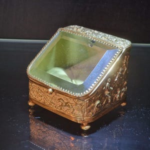 May include: A gold-toned metal display box with a slanted glass lid and a beaded border. The exterior features intricate embossed relief scenes of figures and animals. The box stands on four small ball feet and contains a soft, light-coloured interior.
