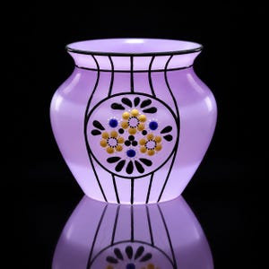 Puede incluir: Un florero de vidrio color violeta claro con un motivo circular central decorado con puntos pintados en amarillo y azul dispuestos en un patrón floral, rodeado de líneas verticales negras que van desde el borde hasta la base del recipiente.