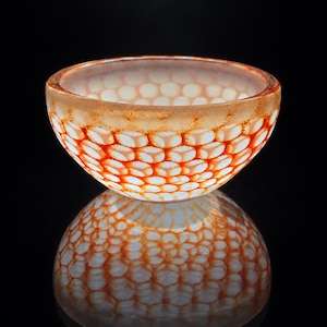 May include: A small, rounded glass bowl featuring a repeating honeycomb pattern in shades of vibrant orange and white. The glass has a translucent quality with a smooth, polished rim and a distinct texture that creates a layered visual effect.