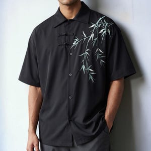 May include: A black short-sleeved button-down shirt featuring a pointed collar and two traditional Chinese-style frog closures at the upper chest. The shirt is decorated with an embroidered design of light green bamboo leaves on the left side.