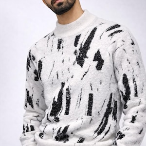 Può includere: Maglione bianco con collo alto e design astratto a pennellate nere. Il maglione ha un aspetto strutturato e dettagli a coste sui polsini e sull'orlo. L'immagine mostra un primo piano del maglione, evidenziando il motivo e il tessuto.