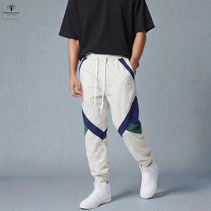 Puede incluir: Pantalones deportivos color crema con detalles geométricos en azul marino y verde. Los pantalones tienen cintura con cordón y puños elásticos. La persona lleva zapatillas blancas y una camiseta negra. Los pantalones son de estilo casual.