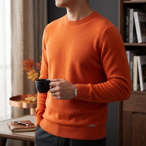 Könnte beinhalten: Ein leuchtend orangefarbener Langarmpullover mit klassischem Rundhalsausschnitt, gerippten Bündchen und einem gerippten Saum. Das Kleidungsstück ist fein gestrickt und verfügt über ein kleines, rechteckiges Metall-Markenetikett am unteren Saum.