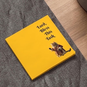 Könnte beinhalten: Ein gelber Haftzettel mit dem Text "Lord, Bless This Task" und einem Katzenbild. Der Zettel liegt auf einer dunkelgrauen Oberfläche, in der Nähe eines Laptops und einer Topfpflanze.