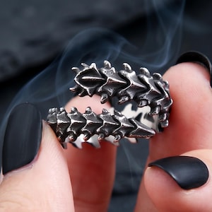 Könnte beinhalten: Ein silberfarbener Metallring mit einem strukturierten, segmentierten Design, das an eine Wirbelsäule oder eine Reihe überlappender Schuppen erinnert. Der Ring ist offen gestaltet, wobei sich die beiden Enden zueinander biegen und eine dunkle, oxidierte Oberfläche aufweisen.