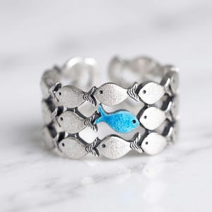 May include: An adjustable silver-toned ring featuring a repeating pattern of small fish silhouettes. The band displays multiple rows of textured silver fish, with one single fish in the centre highlighted in a bright, solid sky blue enamel finish.