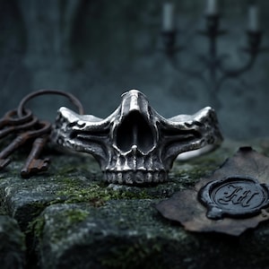 May include: A silver-toned metal ring featuring a detailed skull design with a weathered, dark finish. The skull has prominent eye sockets and a defined jawline, creating a textured appearance. The ring is positioned alongside old keys.