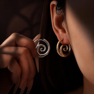 Pode incluir: Um par de brincos de argola com um design de espiral proeminente. Um brinco tem um acabamento em tom prateado polido, enquanto o outro tem um acabamento em tom dourado polido. Ambos os brincos têm formato circular com um padrão central.