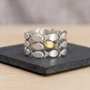 Peut inclure: Une bague large ornée d'un motif répétitif de petits poissons stylisés. La majorité des poissons présente une finition argentée mate, tandis qu'un seul poisson situé au centre du motif arbore une finition dorée contrastante.