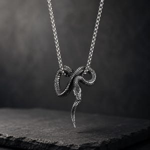 Puede incluir: Un collar de metal plateado con un colgante en forma de serpiente enroscada con escamas detalladas. El colgante cuelga de una cadena de eslabones redondos. La serpiente se muestra en una posición curva de reposo con la cabeza apuntando hacia abajo.