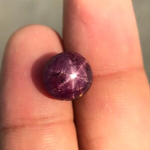Può includere: Una gemma rotonda, di colore viola scuro, con un effetto a stella. La gemma è tenuta tra due dita, mettendo in risalto la sua superficie lucida e le inclusioni naturali. L'effetto a stella è creato dalla riflessione della luce sulla struttura interna della pietra.