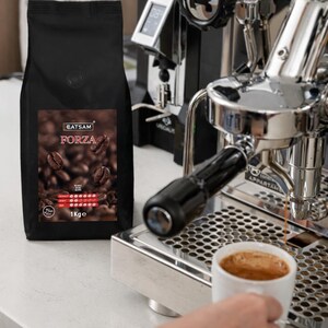 Op de afbeelding: Een zwarte zak EATSAM FORZA koffiebonen met een etiket met koffiebonen. Een witte kop wordt vastgehouden in de buurt van een chromen espressomachine, met koffie die in de kop wordt gegoten. De zak is gelabeld 1Kge.