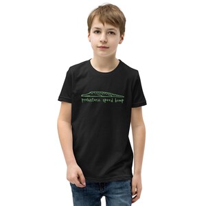 Può includere: T-shirt nera con il contorno verde di un alligatore e la scritta "Prehistoric Speed Bump". La maglietta è realizzata in materiale morbido ed è adatta ai bambini.