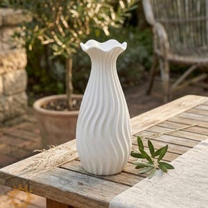 Könnte beinhalten: Eine weiße Keramikvase mit gerüschtem Oberteil und einem wirbelnden, strukturierten Design. Die Vase steht auf einem Holztisch mit einem neutralfarbenen Läufer. Das Bild ist im Freien mit unscharfem Hintergrund aufgenommen.