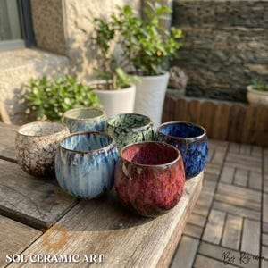 Puede incluir: Una colección de seis tazas de cerámica con forma redondeada y acabado brillante. Las tazas presentan una variedad de colores, incluyendo azul, verde, rojo y marrón. El texto "SOL CERAMIC ART" es visible en la parte inferior de la imagen.