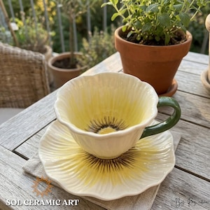 以下が含まれることがあります： 花柄の陶器のティーカップとソーサーのセット。カップとソーサーは白地に黄色と茶色のアクセントがあり、カップには緑色のハンドルが付いています。セットは木製のテーブルの上の灰色のリネンナプキンに置かれています。「SOL CERAMIC ART」の文字が見えます。