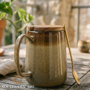 May include: A ceramic mug with a wooden lid and a gold-colored spoon. The mug has a brown and tan gradient design. The handle is curved. The mug is on a wooden surface. The text "SOL CERAMIC ART" is at the bottom.