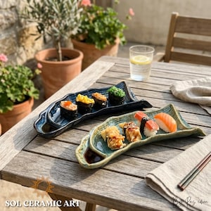 Op de afbeelding: Twee keramische sushi serveerplanken met een golfachtig ontwerp, één blauw en één groen, elk met verschillende sushi rolls. De planken staan op een houten tafel, met een glas water en eetstokjes. De tekst "SOL CERAMIC ART" is zichtbaar.