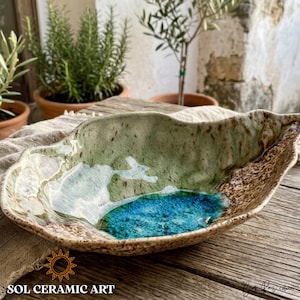 Pode incluir: Uma tigela artesanal de cerâmica em forma de folha com borda marrom salpicada, centro verde e branco e uma piscina azul vibrante. A tigela está sobre uma superfície de madeira rústica, com o texto "SOL CERAMIC ART" na parte inferior.