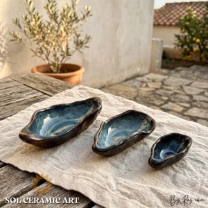 Puede incluir: Tres cuencos de cerámica hechos a mano de diferentes tamaños, con exterior marrón oscuro e interior azul con efecto veteado. Los cuencos se exhiben sobre un paño de color neutro. El texto "SOL CERAMIC ART" es visible.