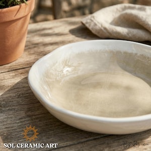 Könnte beinhalten: Eine weiße Keramikschale mit beigem Innenraum, die eine einzigartige, unregelmäßige Form aufweist. Die Schale steht auf einer Holzoberfläche, mit einem Terrakotta-Topf und einem Leinentuch im Hintergrund. Der Text "SOL CERAMIC ART" ist sichtbar.