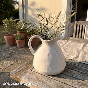 May include: A cream-colored ceramic pitcher with a handle, filled with dried grasses and olive branches. The pitcher sits on a weathered wooden table. The text "SOL CERAMIC ART" is visible at the bottom of the image.