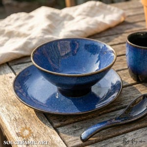 May include: A blue ceramic bowl, saucer, and spoon set with a speckled glaze and a brown rim. The bowl sits on the saucer. The spoon is curved. The set is on a wooden table. The text "SOL CERAMIC ART" is visible.