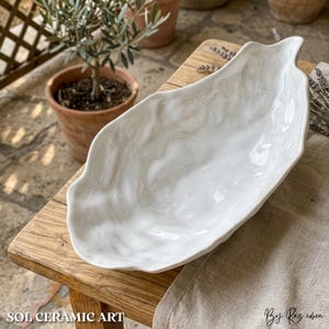 Könnte beinhalten: Eine weiße Keramik-Servierschale in Blattform mit Marmormuster. Die Schale steht auf einer Holzoberfläche. Der Text "SOL CERAMIC ART" befindet sich unten links. Die Signatur des Künstlers befindet sich unten rechts.